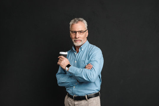 Portrait of adult man 60s with grey hair and beard posing on camera with holding takeaway coffee from paper cup, isolated over black background
