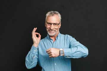 Photo of pleased gentleman 60s with grey hair wearing eyeglasses showing ok sign being on time, isolated over black background