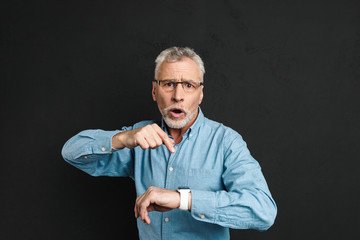 Photo of uptight gentleman 60s with grey hair wearing eyeglasses pointing finger at his wrist watch with shock, isolated over black background