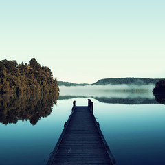 Picture of a minimalist blue landscape of a dock next to a beautiful calm lake. There are some leafy trees and hazy mountains in the the scene. The landscape is reflected on the water.