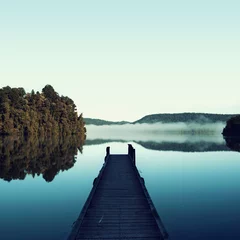 Foto auf Acrylglas Blau Bild einer unbedeutenden blauen Landschaft eines Docks nahe bei einem schönen ruhigen See. Es gibt einige belaubte Bäume und dunstige Berge in der Szene. Die Landschaft spiegelt sich auf dem Wasser.