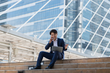 Young Asian businessman happy talking on mobile phone and looking at laptop Sitting in a city Bangkok Thailand. Concept of communication.