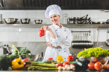 smiling chef holding red bell pepper and looking at camera at restaurant kitchen