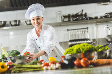 smiling attractive chef taking bowl with vegetables and looking at camera at restaurant kitchen
