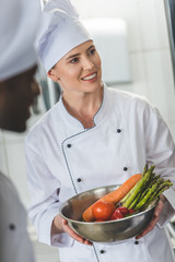 smiling chef holding bowl with vegetables and looking away at restaurant kitchen