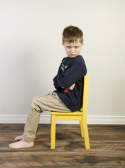 An angry little boy sits in a yellow time out chair on a wooden floor