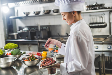 attractive chef adding herbs to raw steaks at restaurant kitchen