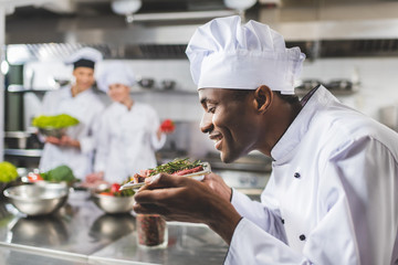 smiling african american chef sniffing raw meat with herbs at restaurant kitchen