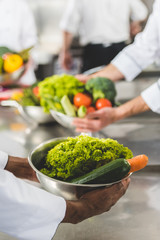 cropped image of multiethnic chefs holding bowls with vegetables at restaurant kitchen
