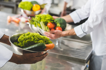 cropped image of multicultural chefs holding bowls with vegetables at restaurant kitchen