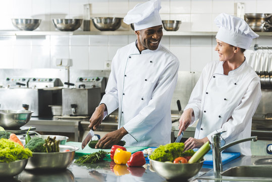 smiling multicultural chefs cutting vegetables at restaurant kitchen and looking at each other