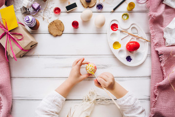 Top view of handmade easter objects laying on white wooden desk, daughter's hands painting, decorate eggs, holding brushed. Day before Easter, child painting eggs for Easter