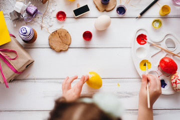 Top view of handmade easter objects laying on white wooden desk, daughter's hands painting, decorate eggs, holding brushed. Day before Easter, child painting eggs for Easter