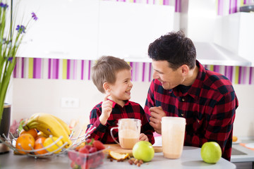 Obraz na płótnie Canvas Young happy father and son in red shirt are tasting freshly made fruit smoothie while laughing and looking at each other at the kitchen counter.