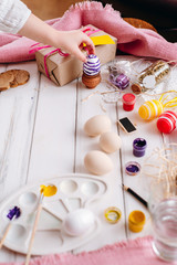 Obraz na płótnie Canvas Top view of handmade easter objects laying on white wooden desk, daughter's hands painting, decorate eggs, holding brushed. Day before Easter, child painting eggs for Easter