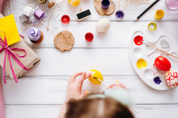 Top view of handmade easter objects laying on white wooden desk, daughter's hands painting, decorate eggs, holding brushed. Day before Easter, child painting eggs for Easter