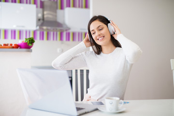 Young relaxed happy girl with headphones on looking at laptop drinking coffee and enjoying music while sitting at the kitchen table.