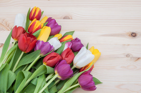 A large bouquet of brightly colored spring tulips in the lower left corner of the image on a background of light wood.