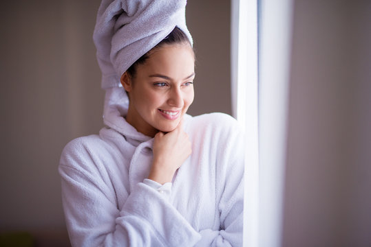 Young Beautiful Joyfull Woman In A Robe With A Towel Around Her Hair Is Smiling And Feeling Fresh After The Shower While Looking Out Of The Window And Feeling Cozy.