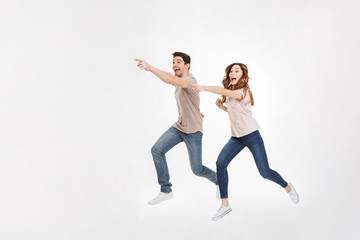Fototapeta na wymiar Full-length picture of delighted couple man and woman in casual t-shirt and jeans running and pointing fingers on copyspace with smile, isolated over white background