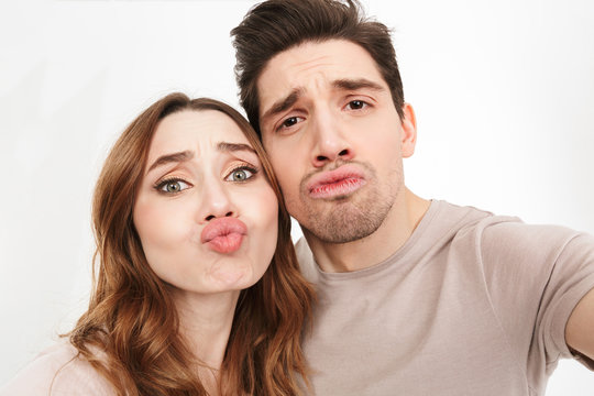 Closeup picture of amusing couple in relationship looking on camera and blowing air kiss while photographing themselves, isolated over white background