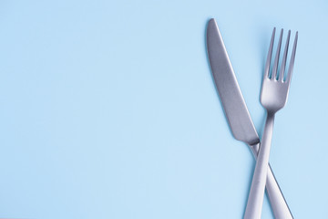 Crossed steel knife and fork on tredny soft blue paper background with side copy space