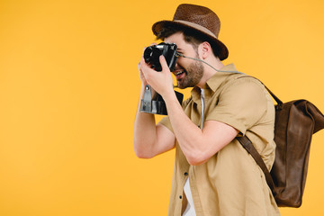 side view of young man in hat with backpack photographing with camera isolated on yellow