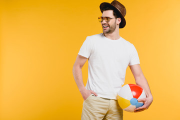 handsome smiling young man in hat and sunglasses holding inflatable ball and looking away isolated on yellow