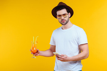 handsome young man looking at camera while holding glass of summer cocktail and using smartphone isolated on yellow - Powered by Adobe