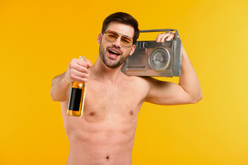 Fototapeta na wymiar young shirtless man in sunglasses holding bottle of summer drink and tape recorder and smiling at camera isolated on yellow