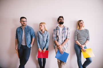 Group of young stylish people leaning against the wall bored before a job interview with folders in...