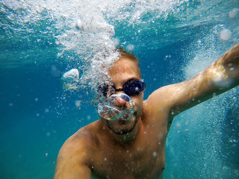 Close up view of young adventuristic bearded man with googles diving in the exotic turquoise sea with a lot of bubbles around him.