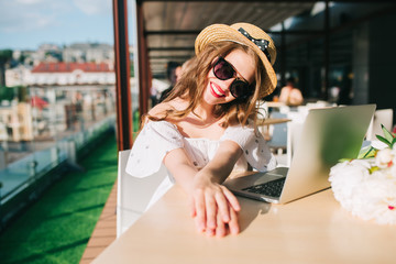 Beautiful girl with long hair in hat sits at  table on the terrace in cafe . She wears a white dress with bare shoulders, red lipstick, sunglasses . She is stretching hands smiling at the camera .