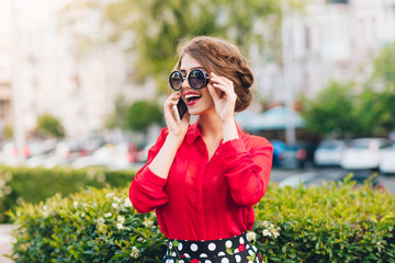 Horizontal portrait of pretty girl in sunglasses walking in park. She wears red blouse and nice hairstyle. She is speaking on phone.