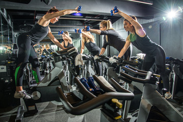 Group of sporty girlfriends training together with dumbbells on cycling class at gym.