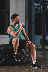 Fototapeta na wymiar sportsman sitting on tire and drinking water after cross training in gym