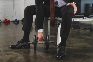 cropped view of sporty businessman in suit training with dumbbell in gym