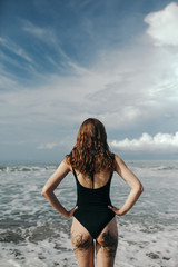 Woman on beach, rear view of beautiful girl with sandy butt wearing black bikini standing back, beautiful sky