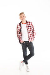 Fototapeta premium Full length portrait of happy educated teenage boy wearing plaid shirt smiling and looking on camera keeping hands in pockets, isolated over white background