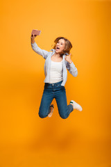 Fototapeta na wymiar Studio portrait of joyful woman wearing jeans and sneakers smiling and showing thumb up while taking selfie with cell phone, isolated over yellow background
