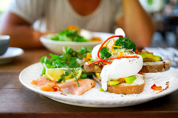 A white plate on a wooden table in a cafe in Bali in Ubud, Canggu. Breakfast in the restaurant with eggs poached, salmon, avocado, beans, salad, red pepper, mushrooms. Blurred background with man