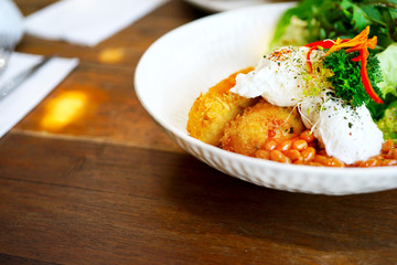 A white plate on a wooden table in a cafe in Bali in Ubud, Canggu. Breakfast in the restaurant with eggs poached, potato croquettes, beans, salad, red pepper, mushrooms. Side view with copy space