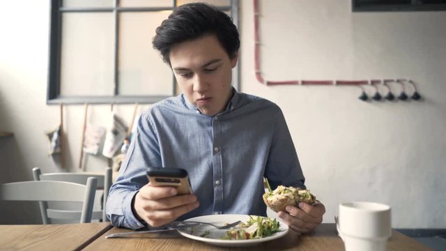 Young teenager with smartphone eating hamburger sitting in cafe
