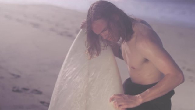 Panning shot of a young man waxing his surfboard at a beach