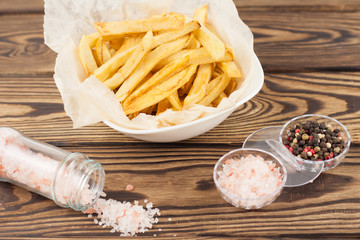 Tasty sticks of french fries in white ceramic bowl with crumpled paper and salt and pepper on old brown rustic wooden table