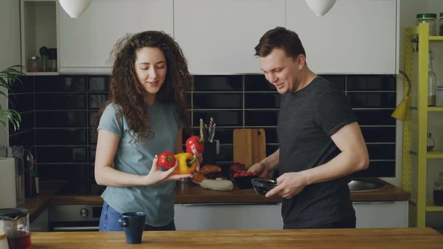 young caucasian loving couple kissing and putting different vegetables and fruits on table to cut them, man giving knife to woman, they are nice and positive