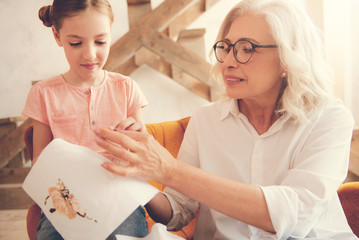 Between generations. Nice pleasant smart woman sitting together with her granddaughter and smiling while teaching her to embroider