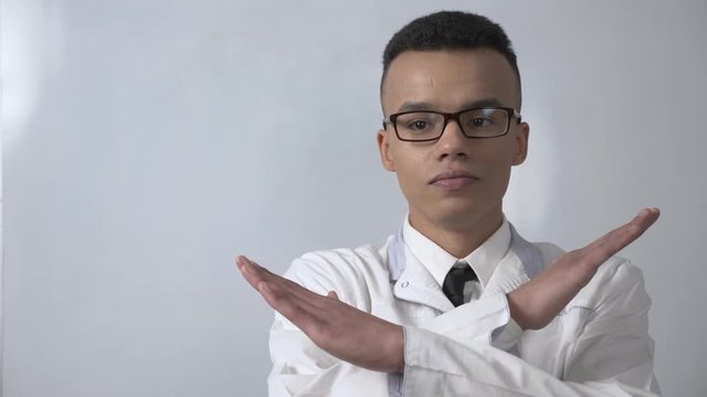 Young successful African scientist in a white coat and glasses showing stop sign, no, never gesture, crossed his arms, looking at the camera, portrait concept. 60 fps