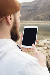 Close-up of a horde in a brown cap in the open air holds a white tablet pc in his hands. A bearded...
