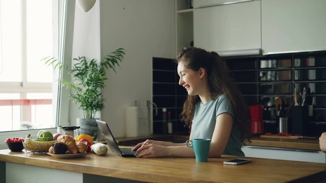 Young curly pretty positive caucasian woman sitting at table and have video call on laptop computer, she sitting in kitchen near window, her boyfriend comes and they skype together in positive way